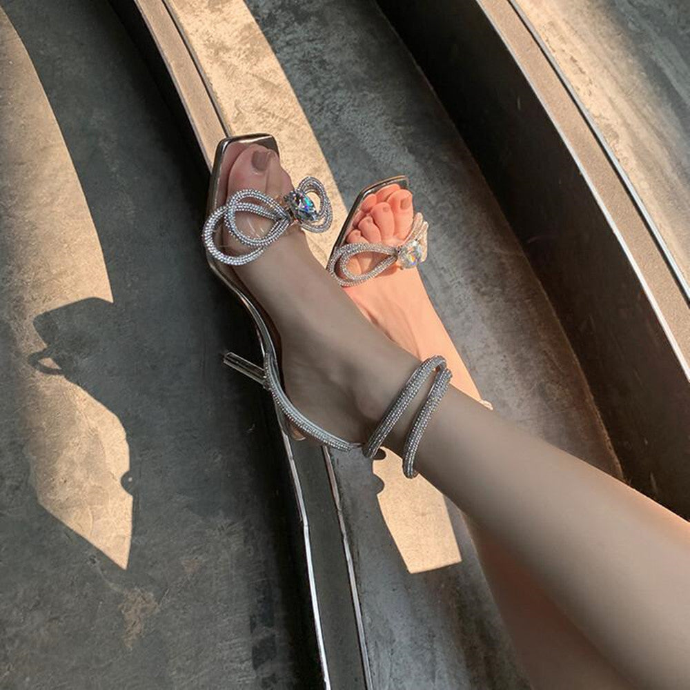 حذاء شفاف بكعب عالٍ مزين بفيونكة مرصعة بأحجار الراين وحزام للكاحل